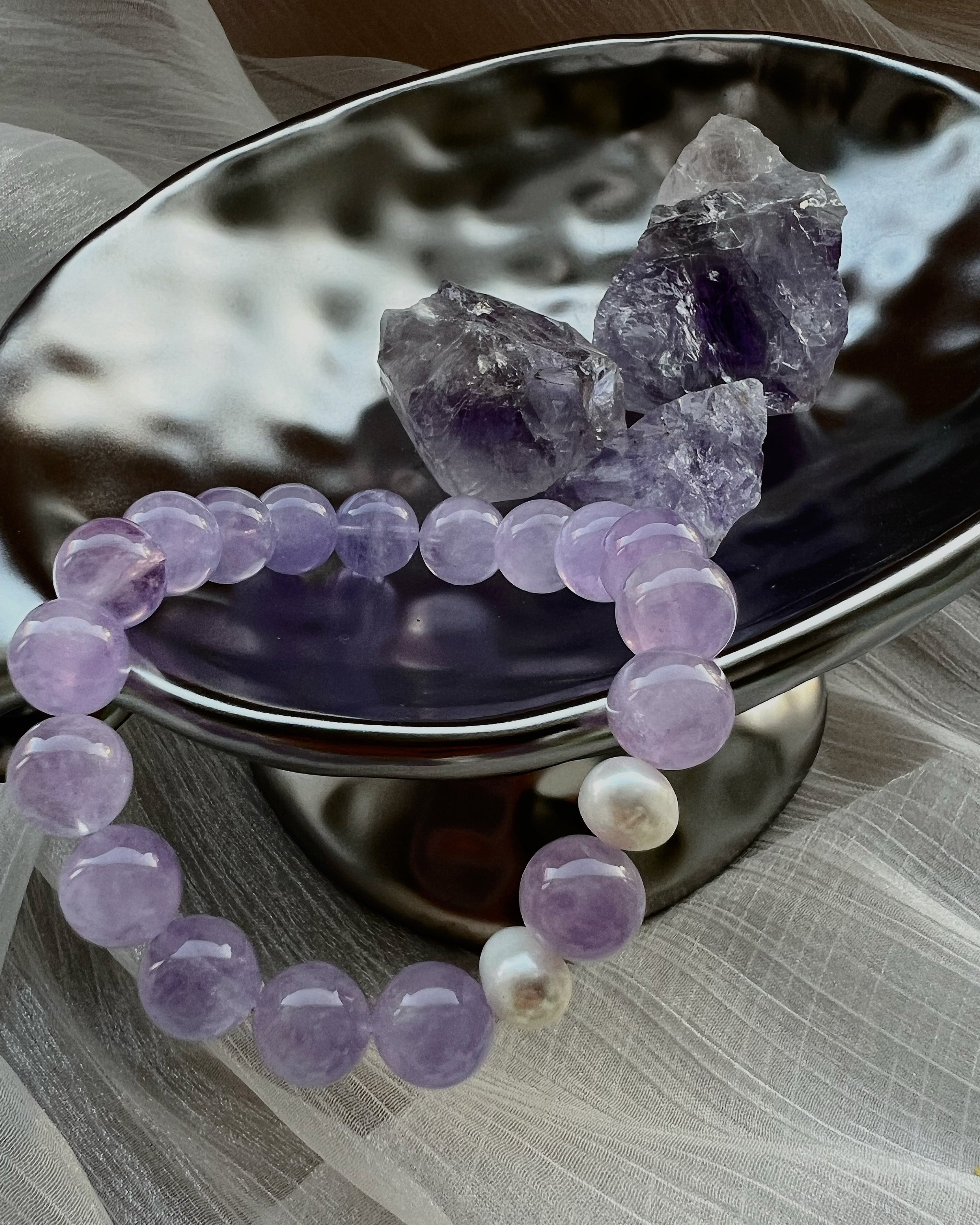 Amethyst crystal bracelet with freshwater pearls displayed on a silver dish alongside raw Amethyst stones – handmade crystal jewelry for balance and healing.