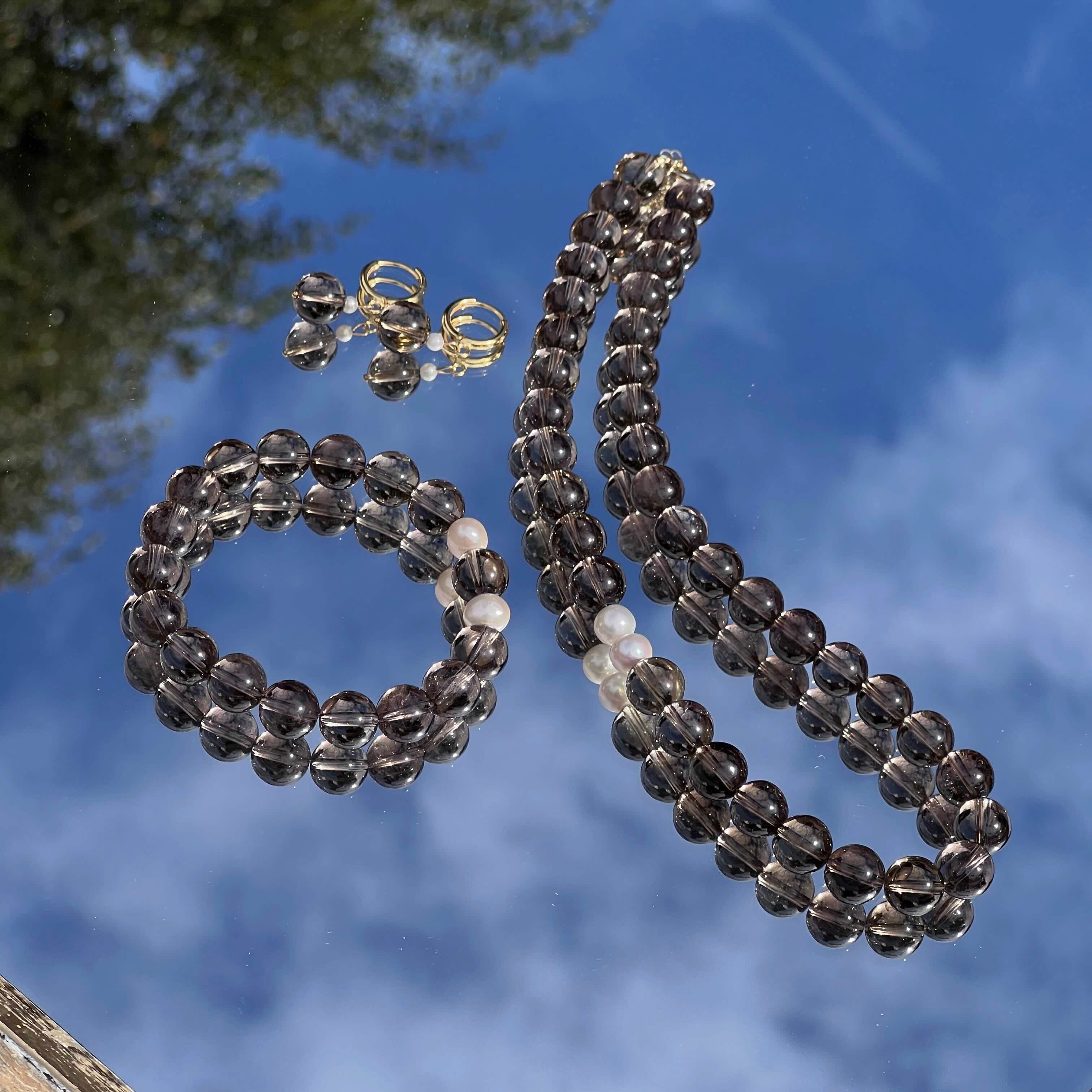 Smoky Quartz bracelet, necklace, and earrings set displayed on a reflective surface under a blue sky. The natural smoky brown crystals are paired with freshwater pearls and gold details, symbolizing grounding, protection, and calm energy.