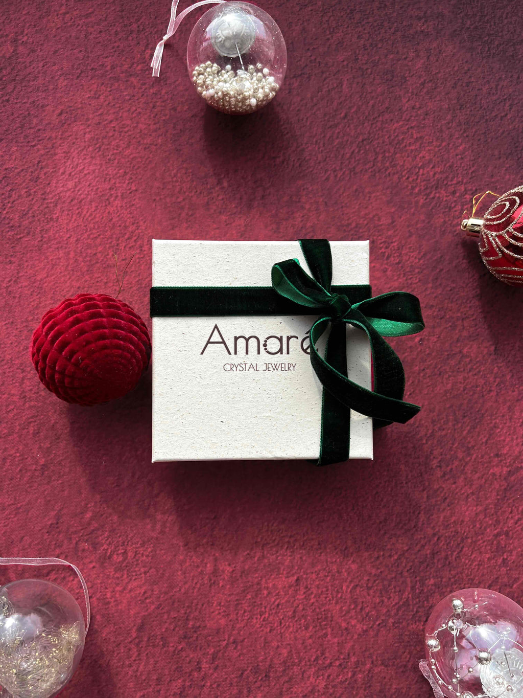 White jewelry box with a green ribbon on a red textured surface with Christmas ornaments.