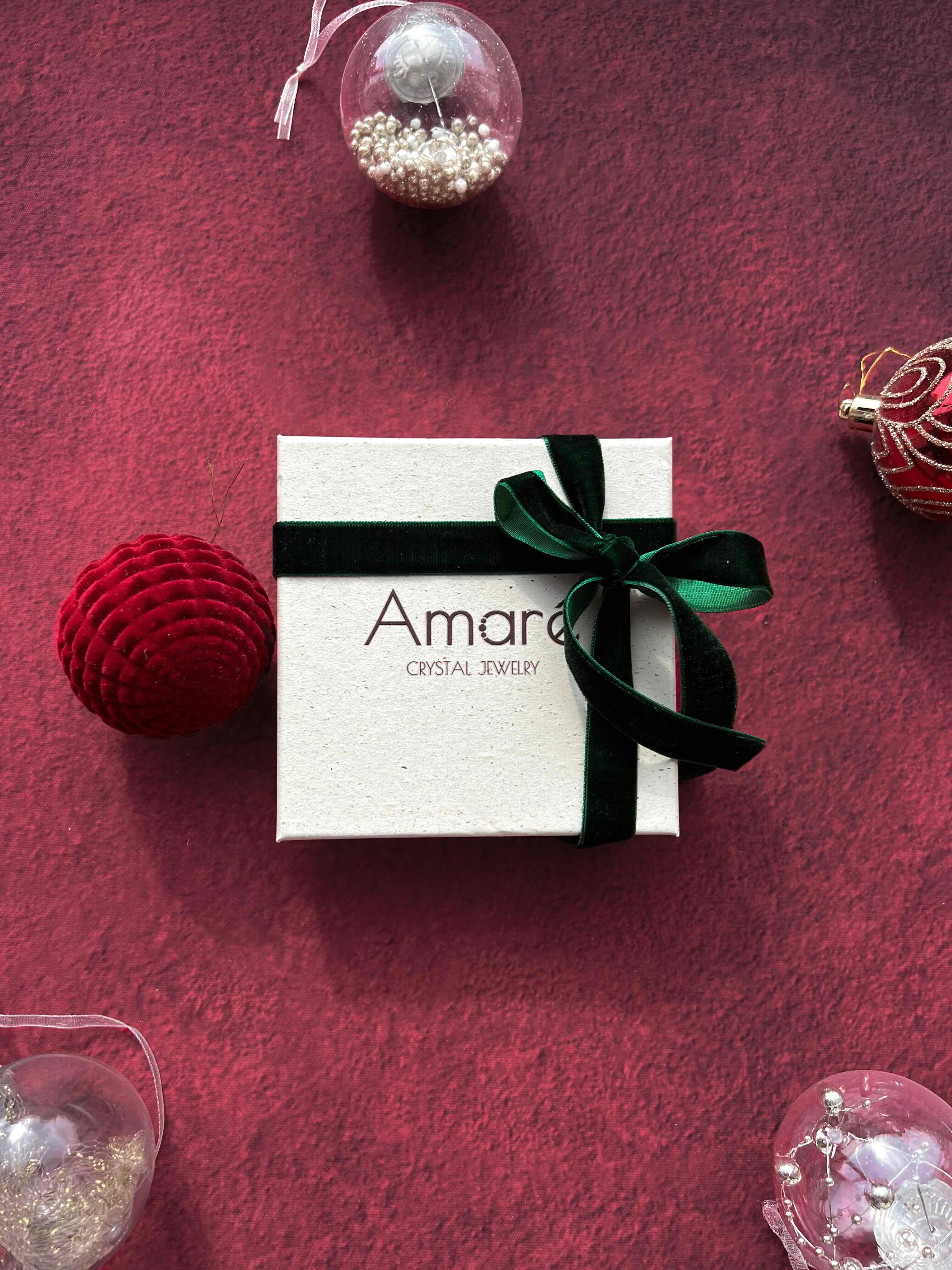 White jewelry box with a green ribbon on a red textured surface with Christmas ornaments.