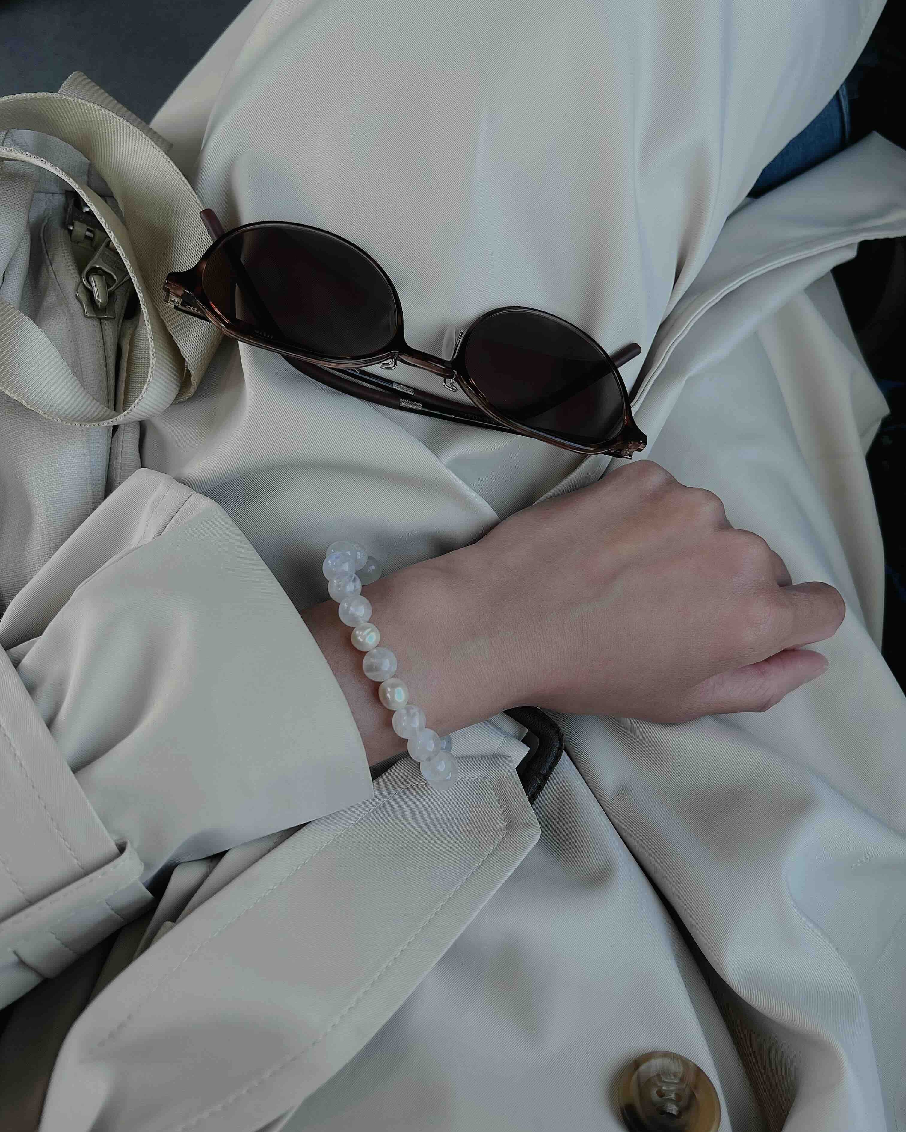 Hand wearing Amaré Blue Moonstone Bracelet styled with a beige trench coat and sunglasses, showcasing the crystal’s soft glow and elegant design.